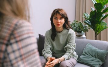 Terapia: o que é, benefícios e por que você deveria investir no seu bem-estar Mulher de moletom verde claro, sentada em um sofá cinza de frente para uma terapeuta