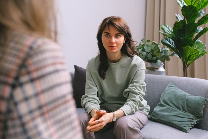 Mulher de moletom verde claro, sentada em um sofá cinza de frente para uma terapeuta