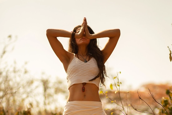 Mulher meditando ativamente em campo aberto, com as palmas das mãos unidas na altura de seu rosto, coluna ereta - transparece paz.