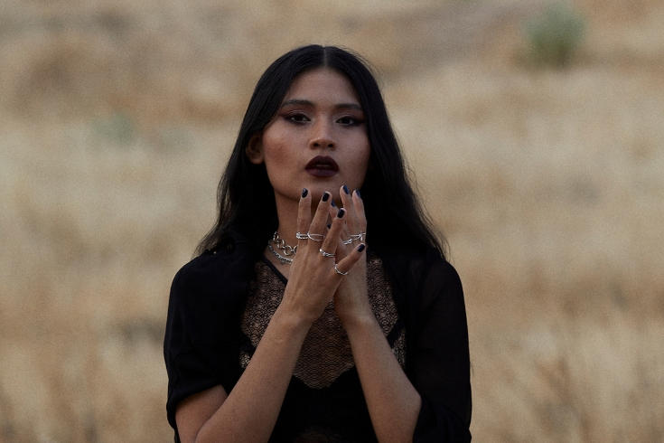 Uma bruxa, vestida de preto, com maquiagem escura, tocando o rosto em um campo aberto.