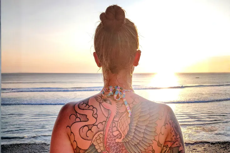 Mulher na praia, com uma grande tatuagem em suas costas, observando o nascer do sol